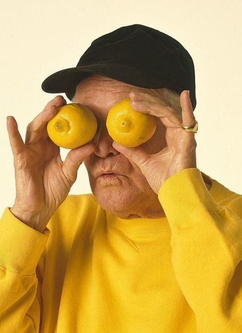 An elderly person in a yellow sweater and black cap playfully holding two lemons over their eyes against a plain background.