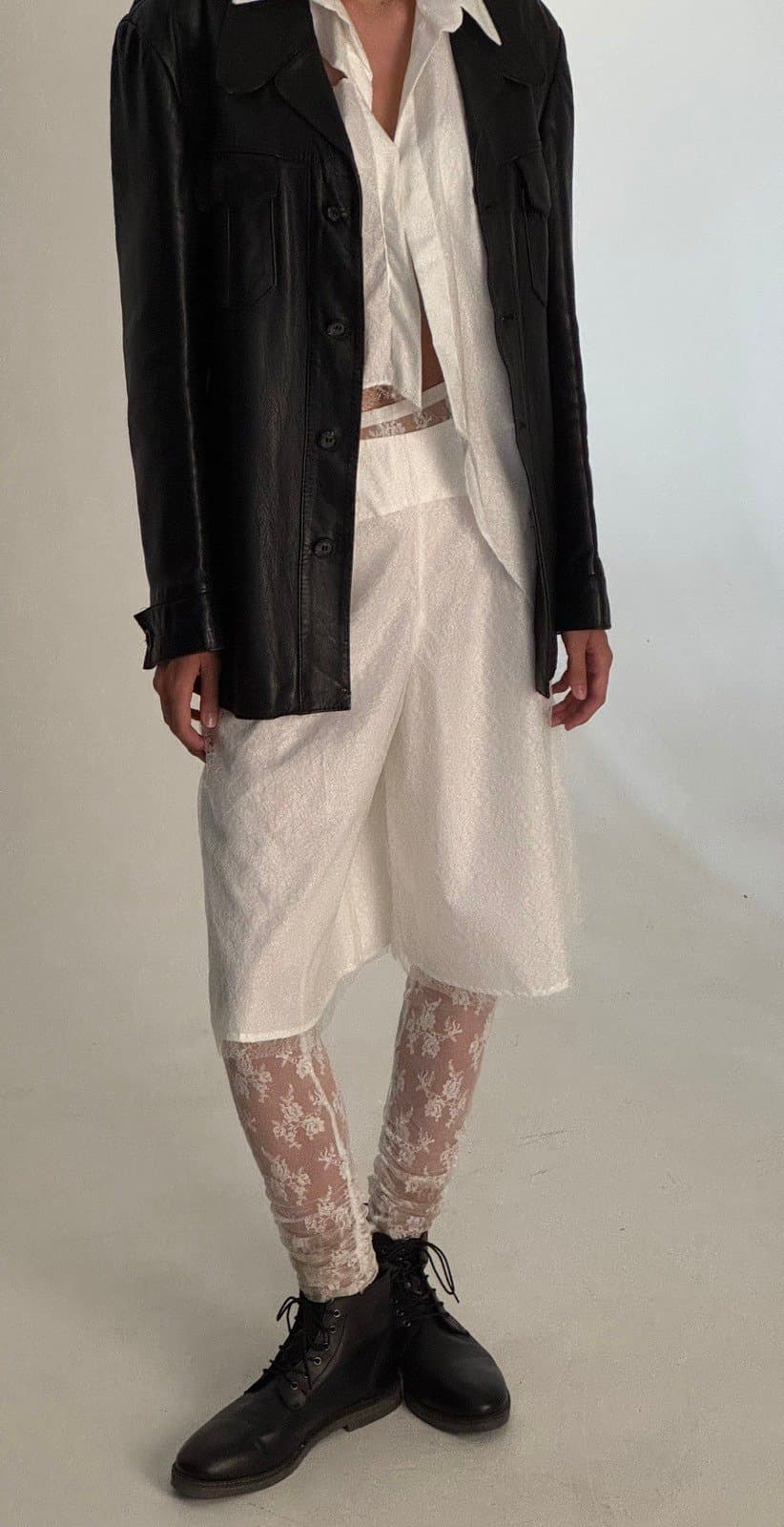 Model in a layered outfit consisting of a long white shirt or dress, black jacket, and lace-printed tights with black shoes.