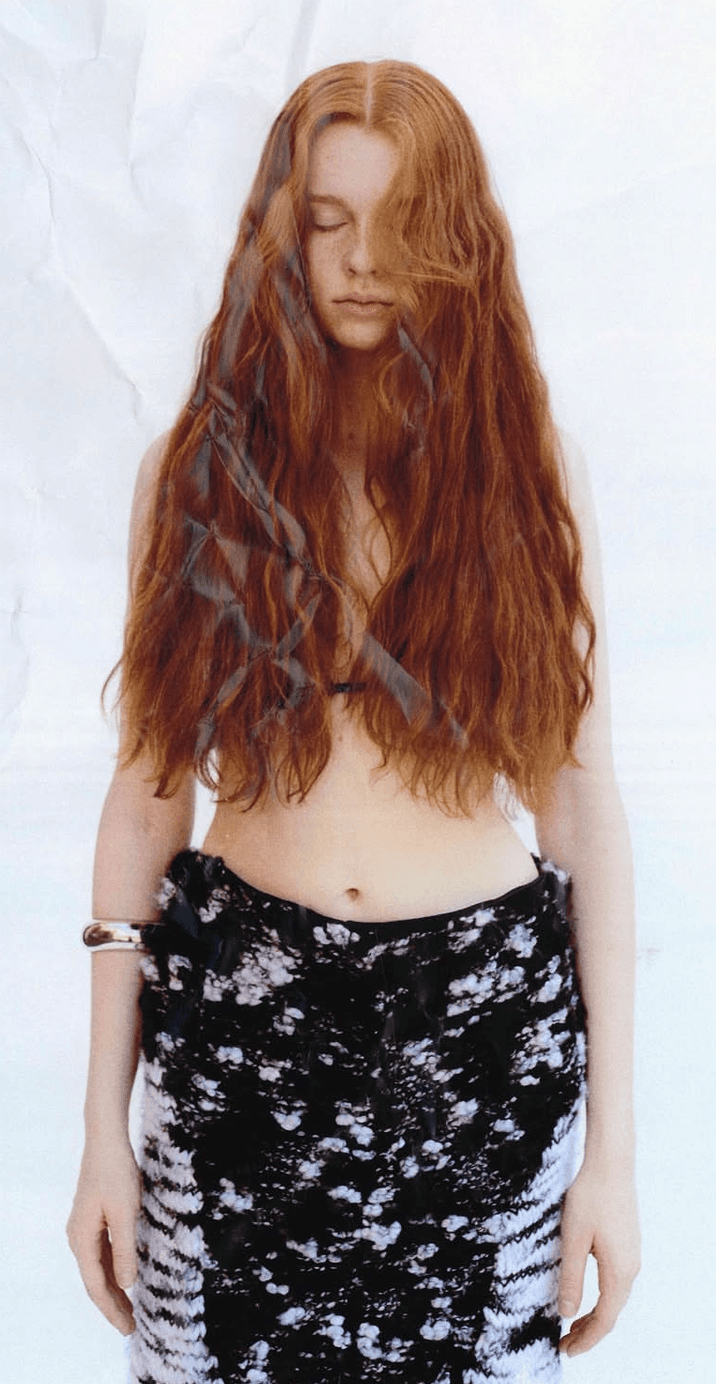 A model with long red hair standing against a white backdrop, wearing a black and white skirt.