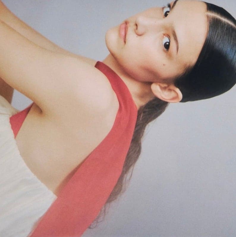 A close-up of a model in a sleeveless white and red garment, looking at the camera with a soft expression.