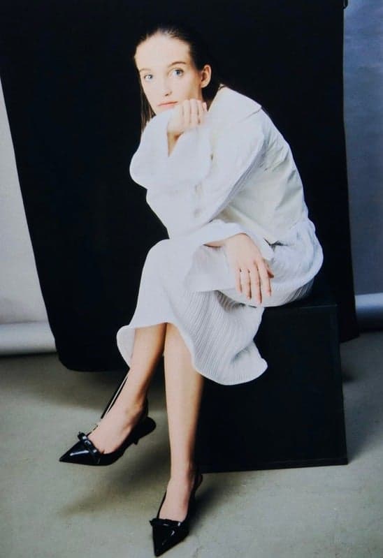 A woman sitting against a dark background, dressed in a white outfit with black pointed shoes, gazing thoughtfully at the camera.