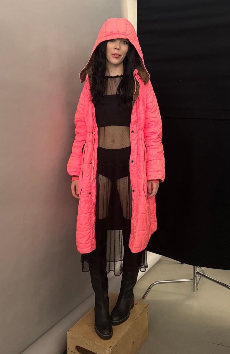 Model wearing a bright pink puffer coat with a hood over a black crop top and leather pants, standing in front of a plain backdrop.