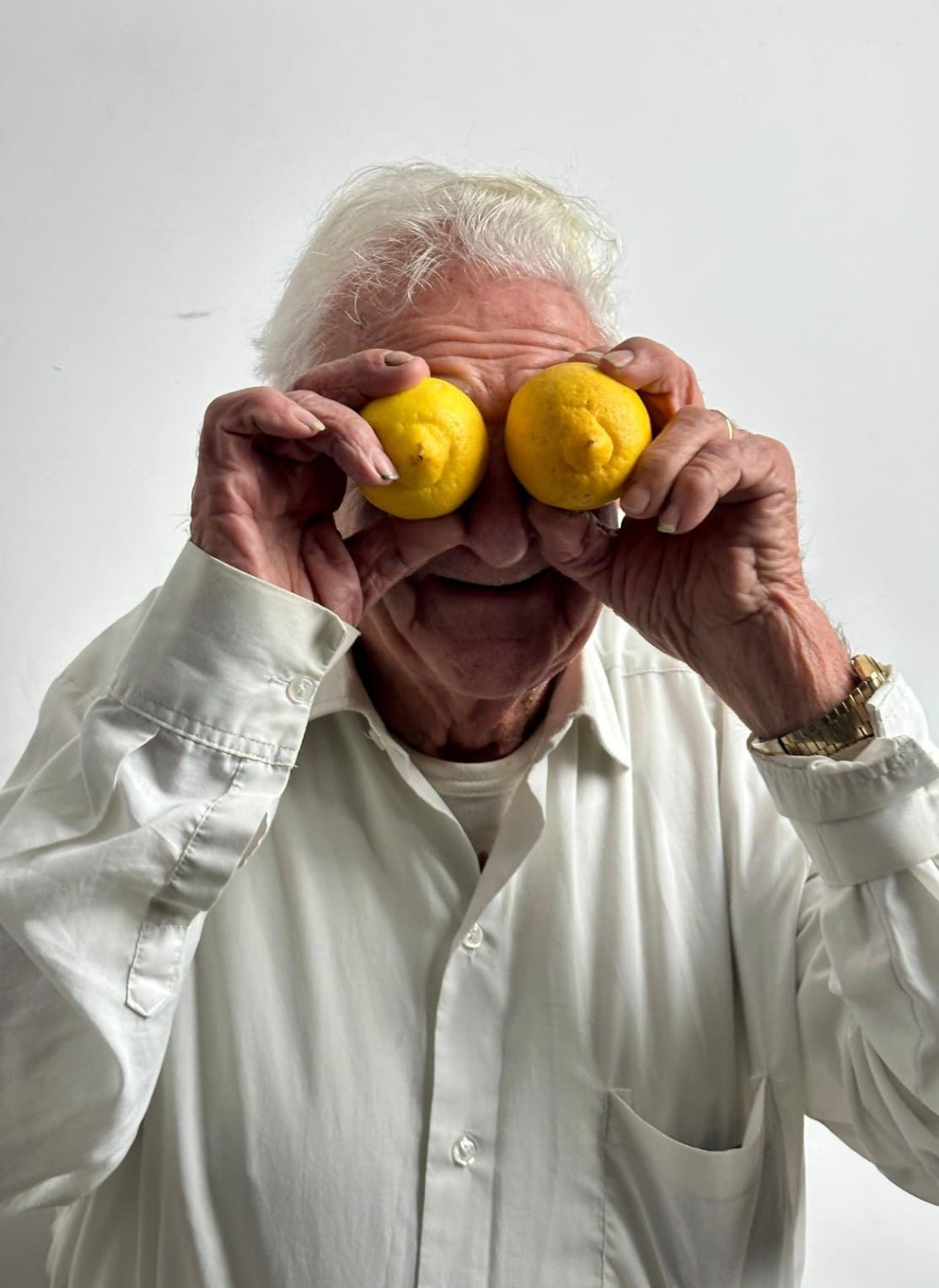 An elderly person with white hair and a white shirt holding two lemons over their eyes and smiling against a light background.
