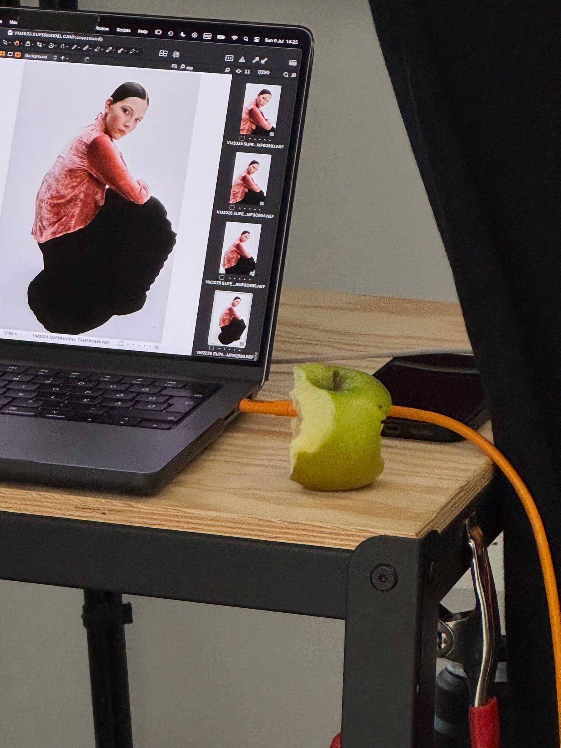 Behind-the-scenes photo showing a laptop displaying a photoshoot image of a model in a red and black outfit, next to an apple and camera equipment on a studio table.